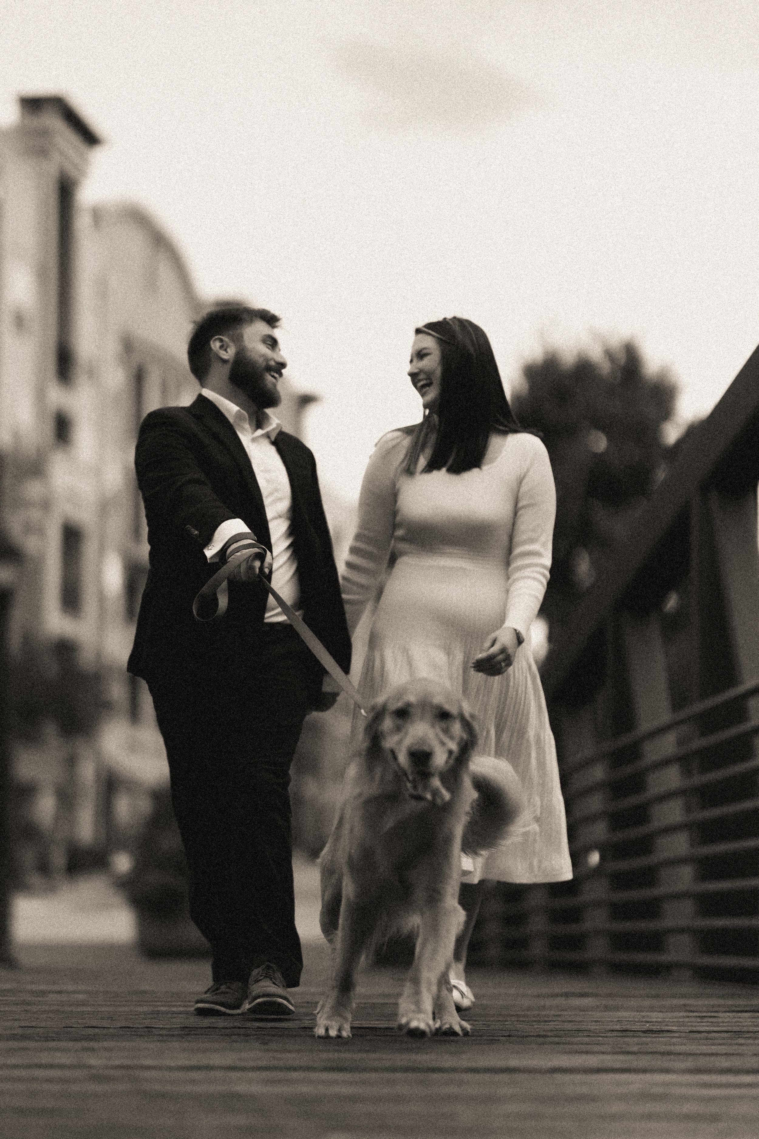 A couple stands on a wooden boardwalk with their fluffy dog in a romantic black and white photo series.