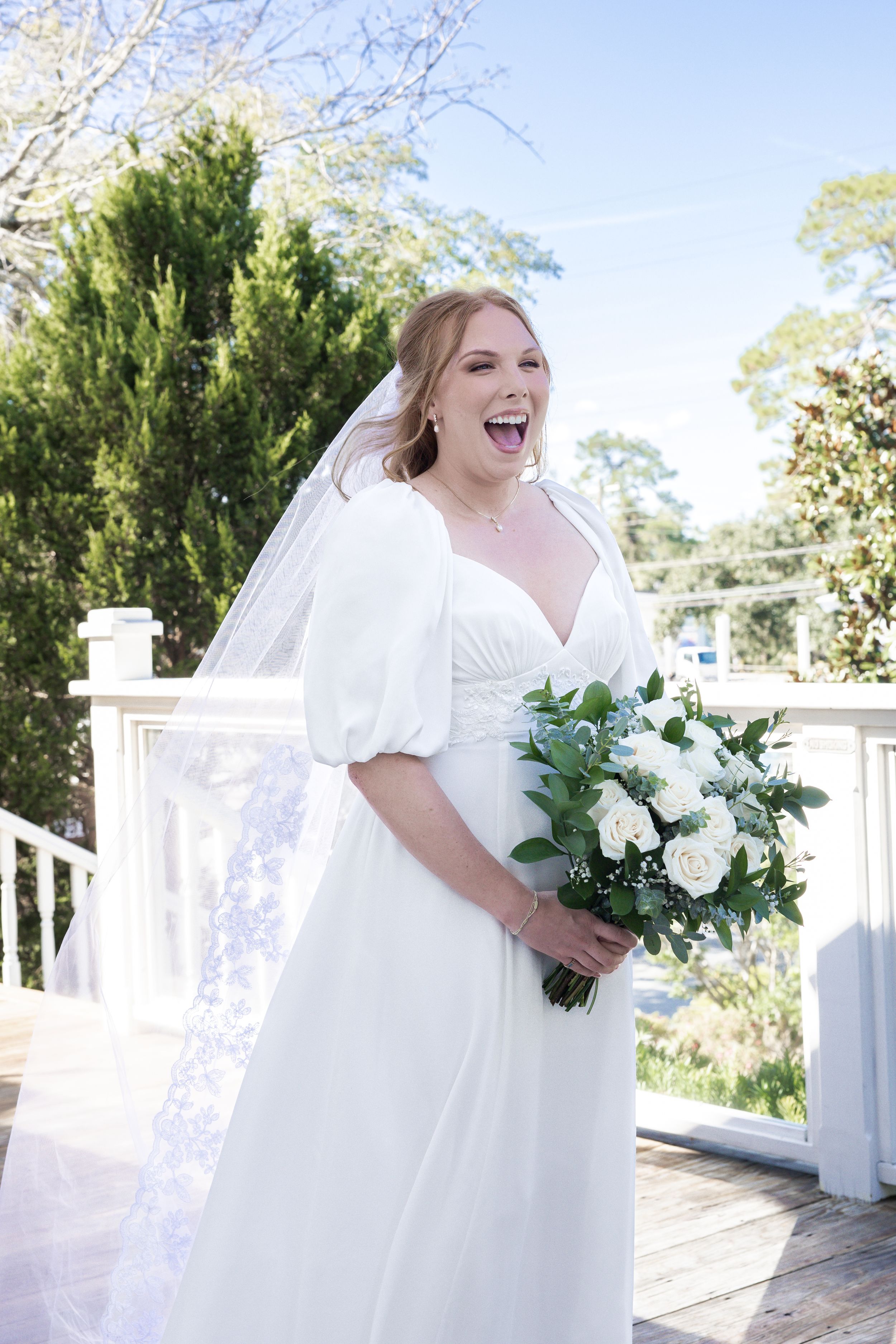 Joyful bride in white puff-sleeve gown holding white rose bouquet, laughing on outdoor deck with veil flowing.