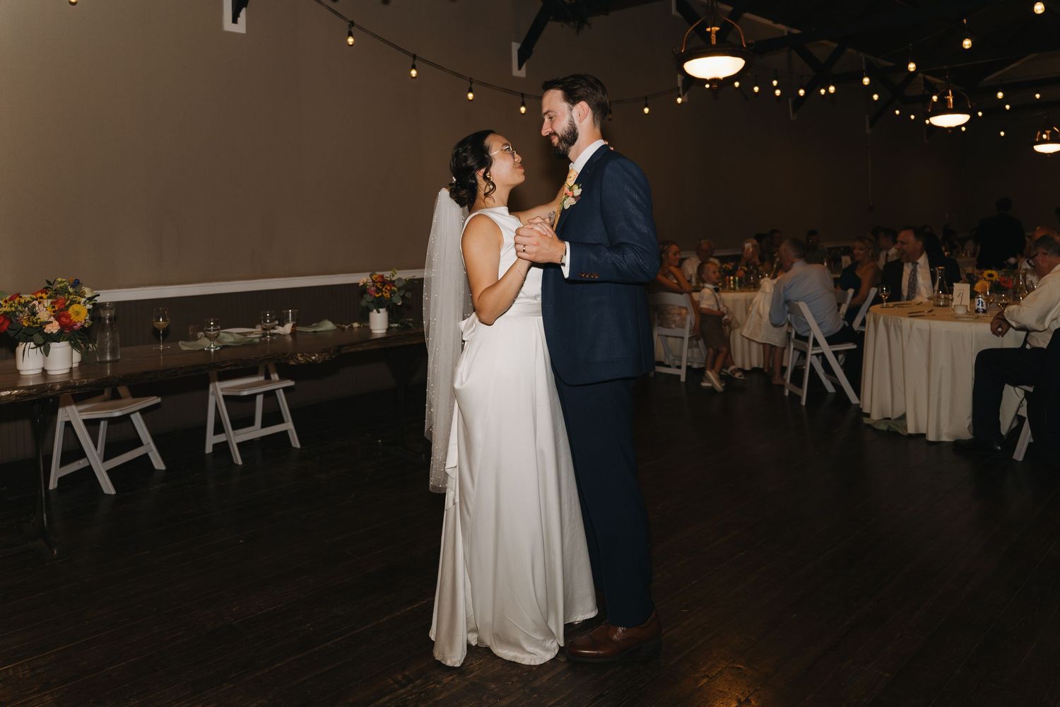 A couple shares their first dance at their wedding reception in a dimly lit venue with string lights.