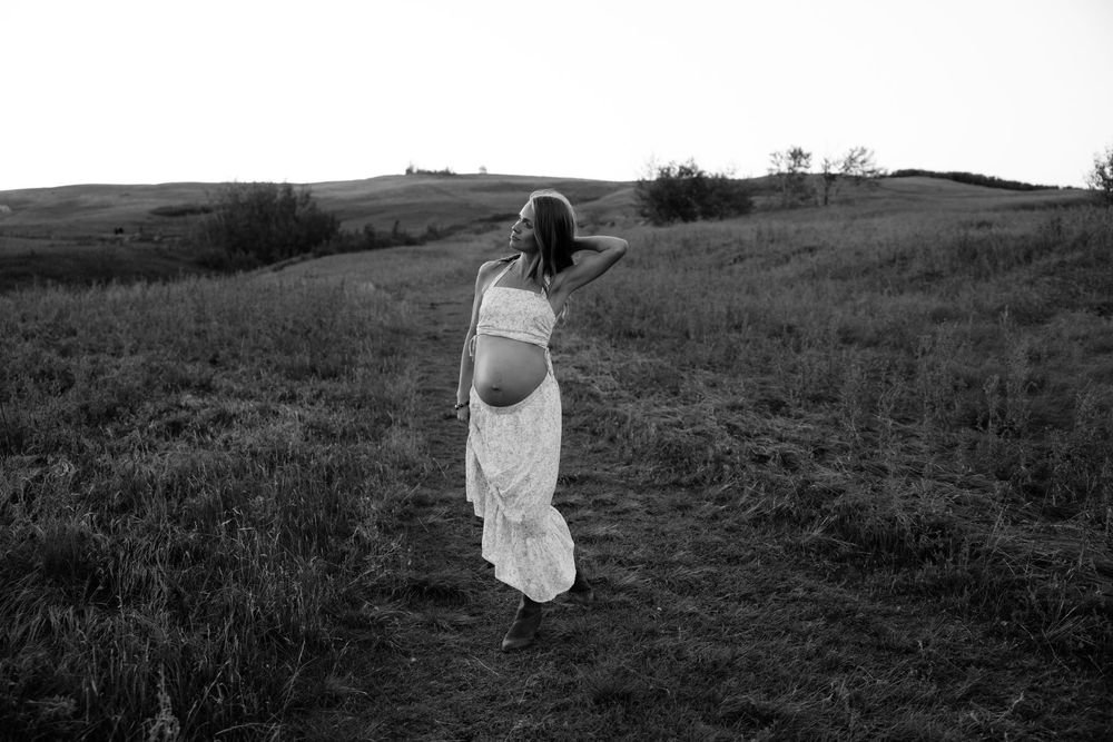 A black and white photograph of a woman in a flowing white dress walking through a grassy hillside meadow.