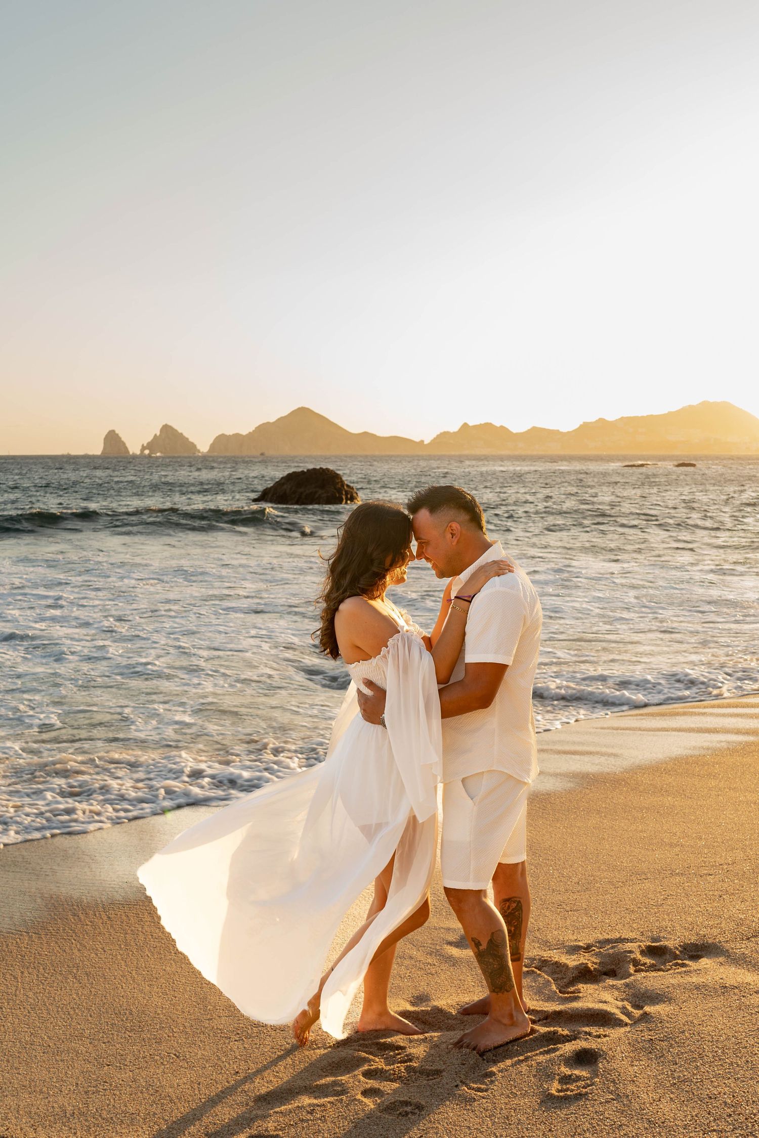 Romantic beach photo of couple in white clothing during golden hour with waves crashing behind.