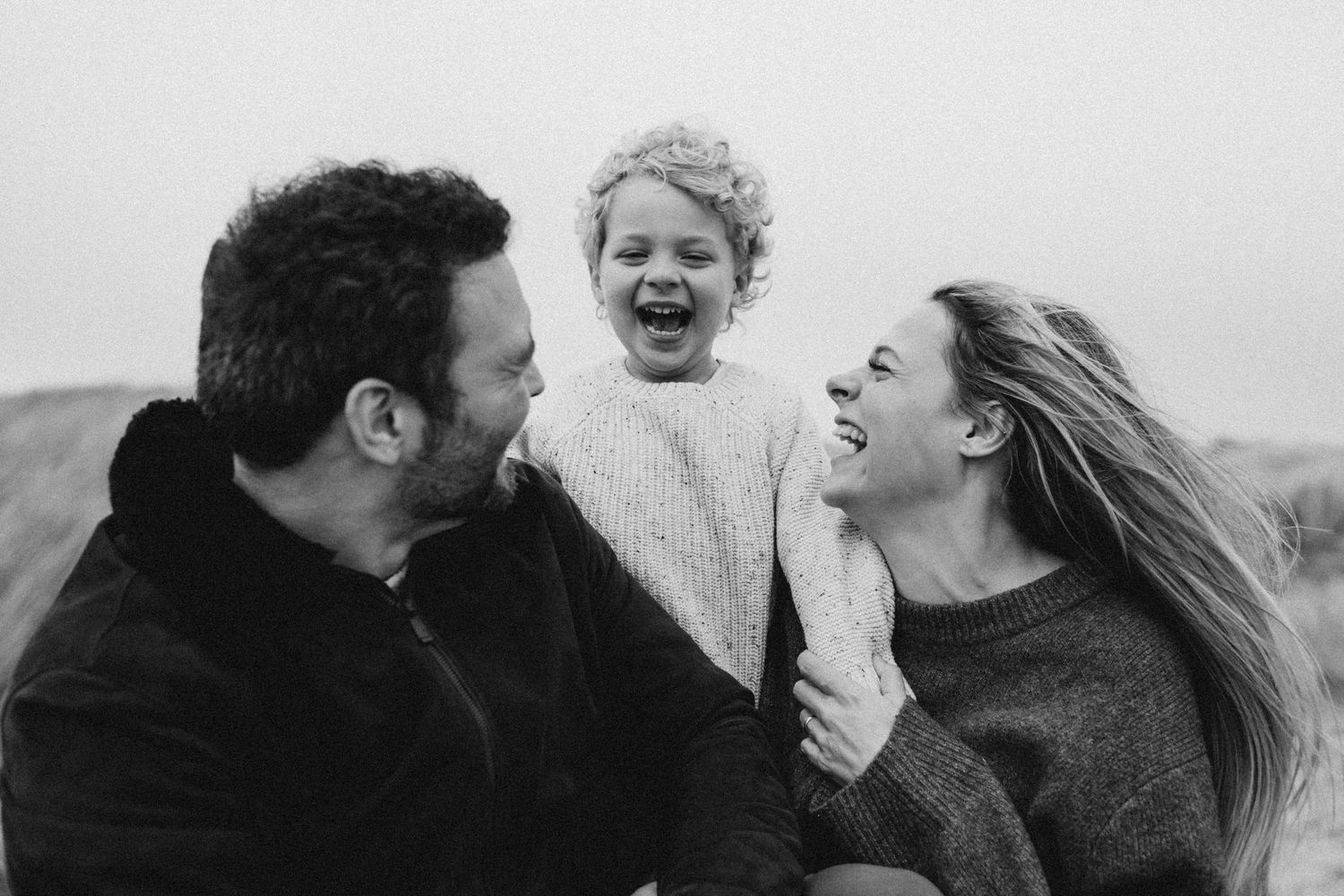 A family of three laughing while the little boy is playing hide and seek behind his parents during a family photo session at the beach Kent