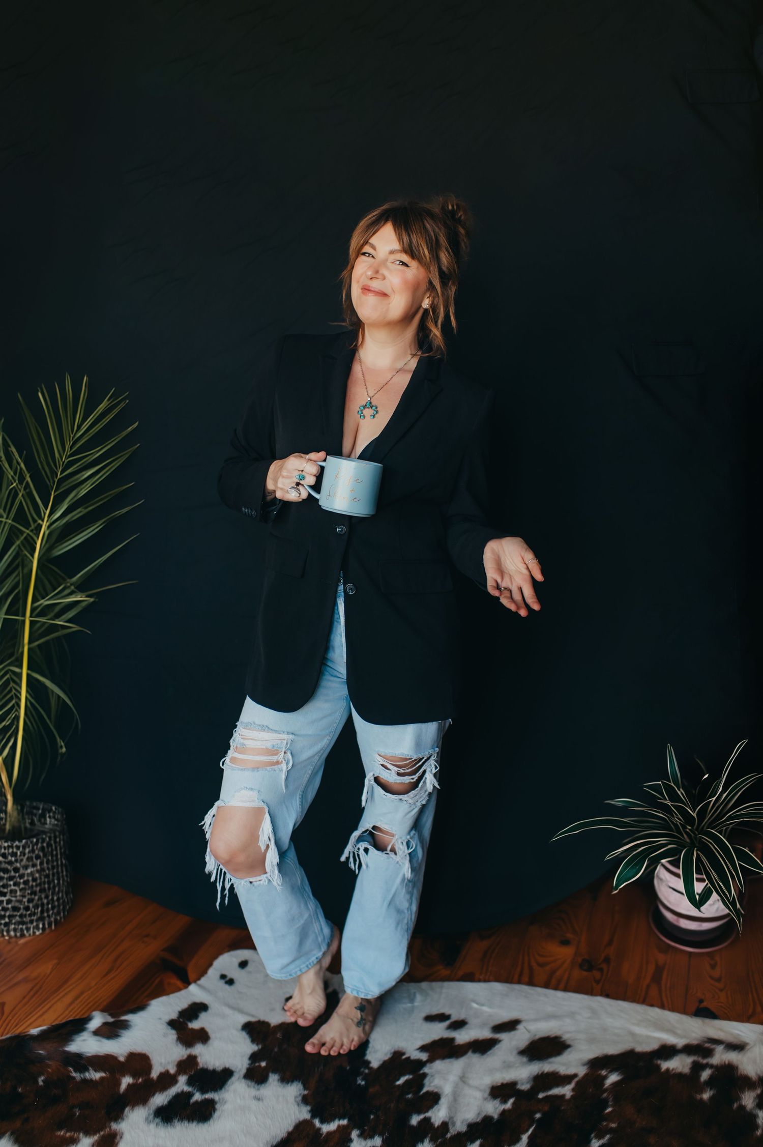 Casual portrait against dark wall wearing blazer and distressed jeans while holding coffee cup.