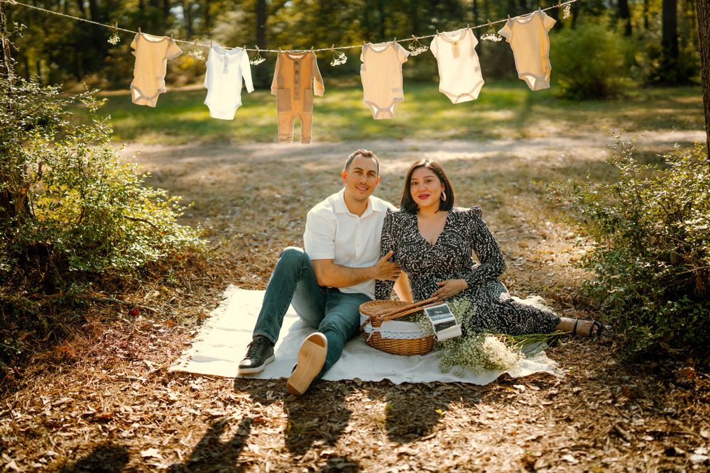 A couple sits on a blanket for a picnic in the woods with baby clothes hanging on a clothesline above them.