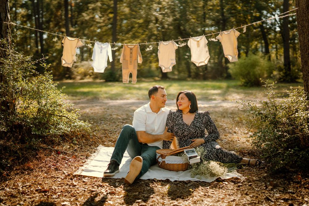 A couple sits on a picnic blanket in the woods with baby clothes hanging on a clothesline behind them.