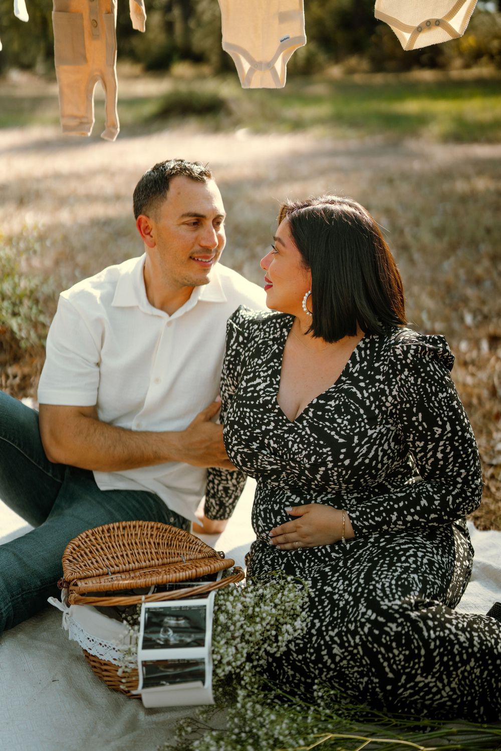 Couple sits on blanket with picnic basket and baby onesies hanging above in golden sunlight.