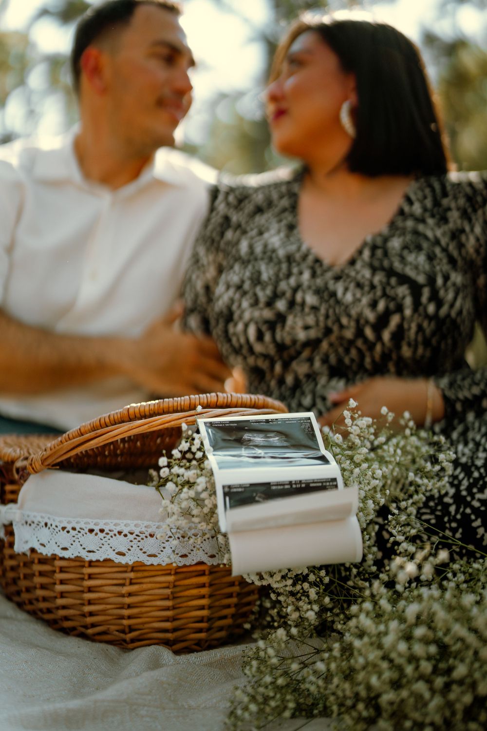 Wicker picnic basket with white lace trim and baby's breath flowers sits in foreground of romantic outdoor setting.