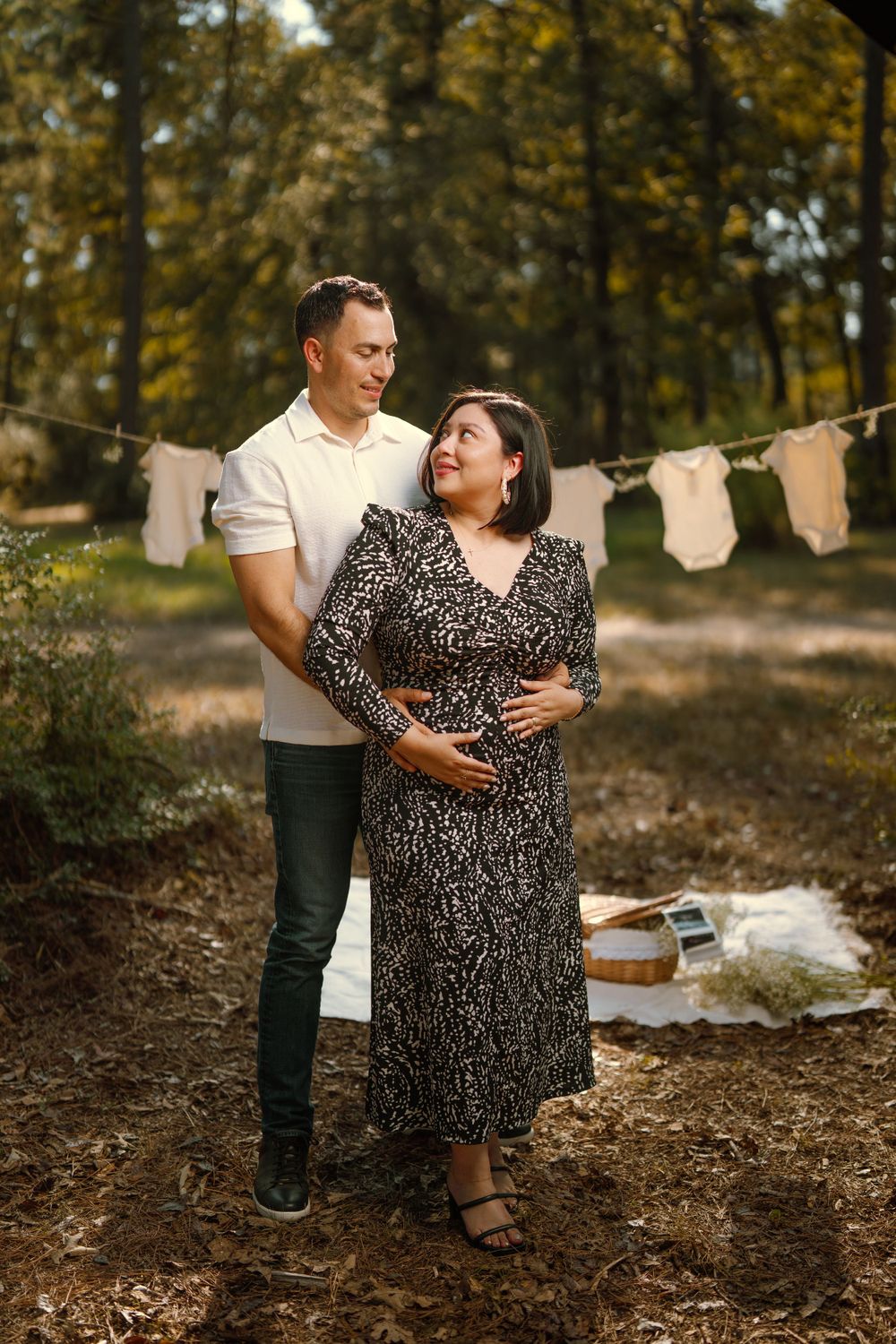 Expecting couple poses outdoors in autumn forest setting with baby clothes hanging on clothesline in background.