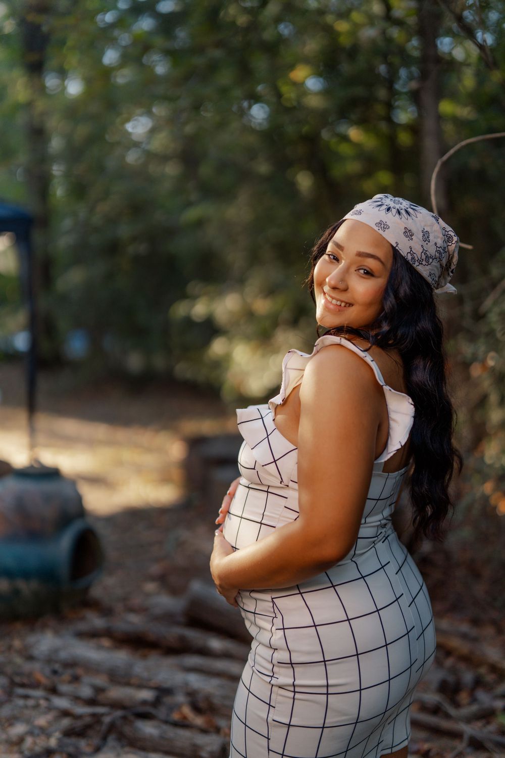 Expectant mother in white gridded dress and floral headband poses outdoors with natural bokeh background.