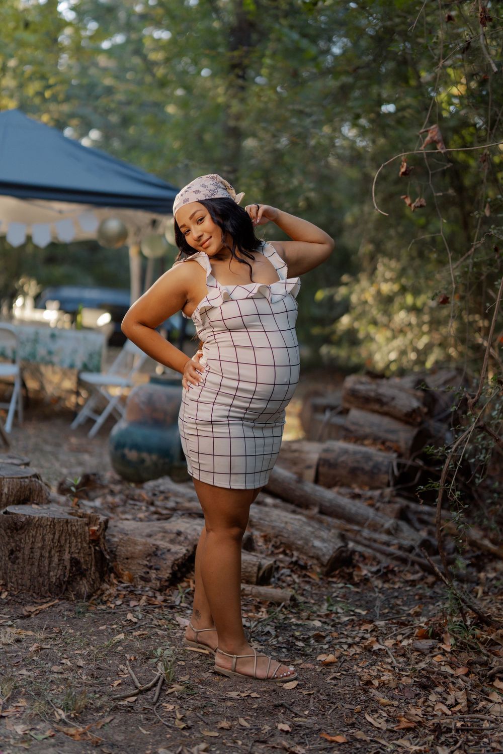 A pregnant woman in a checkered mini dress poses outdoors at sunset among trees and fallen logs.
