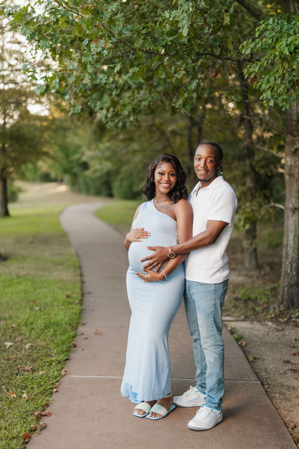 Expectant couple embraces on a tree-lined path wearing light blue dress and white shirt during outdoor maternity photo session.