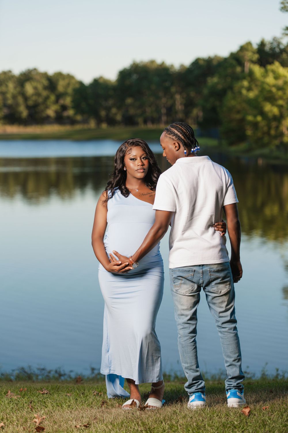 A couple stands together by a serene lake, with the person in white dress and other in casual attire against nature backdrop.