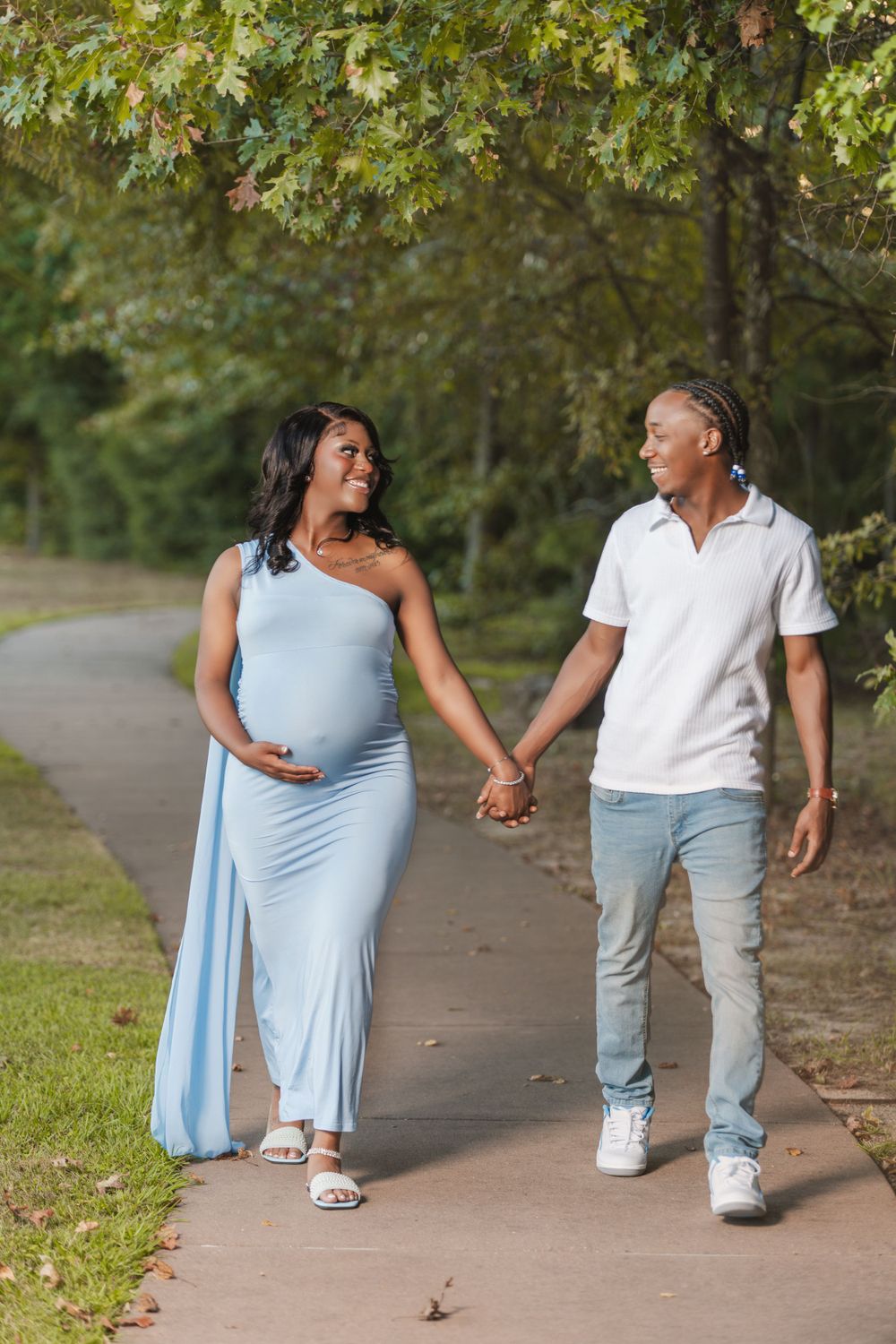 Couple walking hand in hand on a tree-lined path, with one wearing a flowing light blue dress on a sunny day.