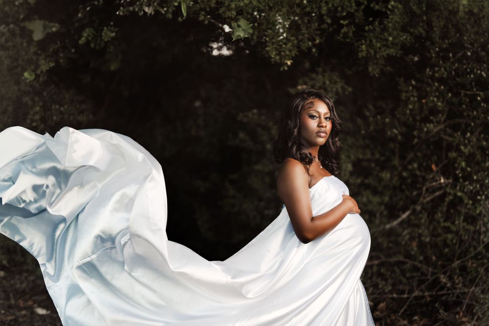 A flowing white maternity dress billows dramatically in the wind against a dark forest backdrop during an outdoor photo session.