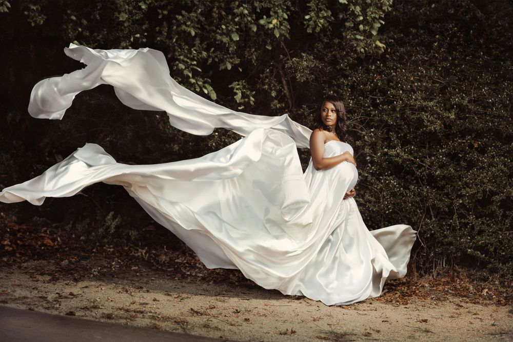 A flowing white wedding dress billows dramatically in the wind against a dark forest backdrop on a dirt path.
