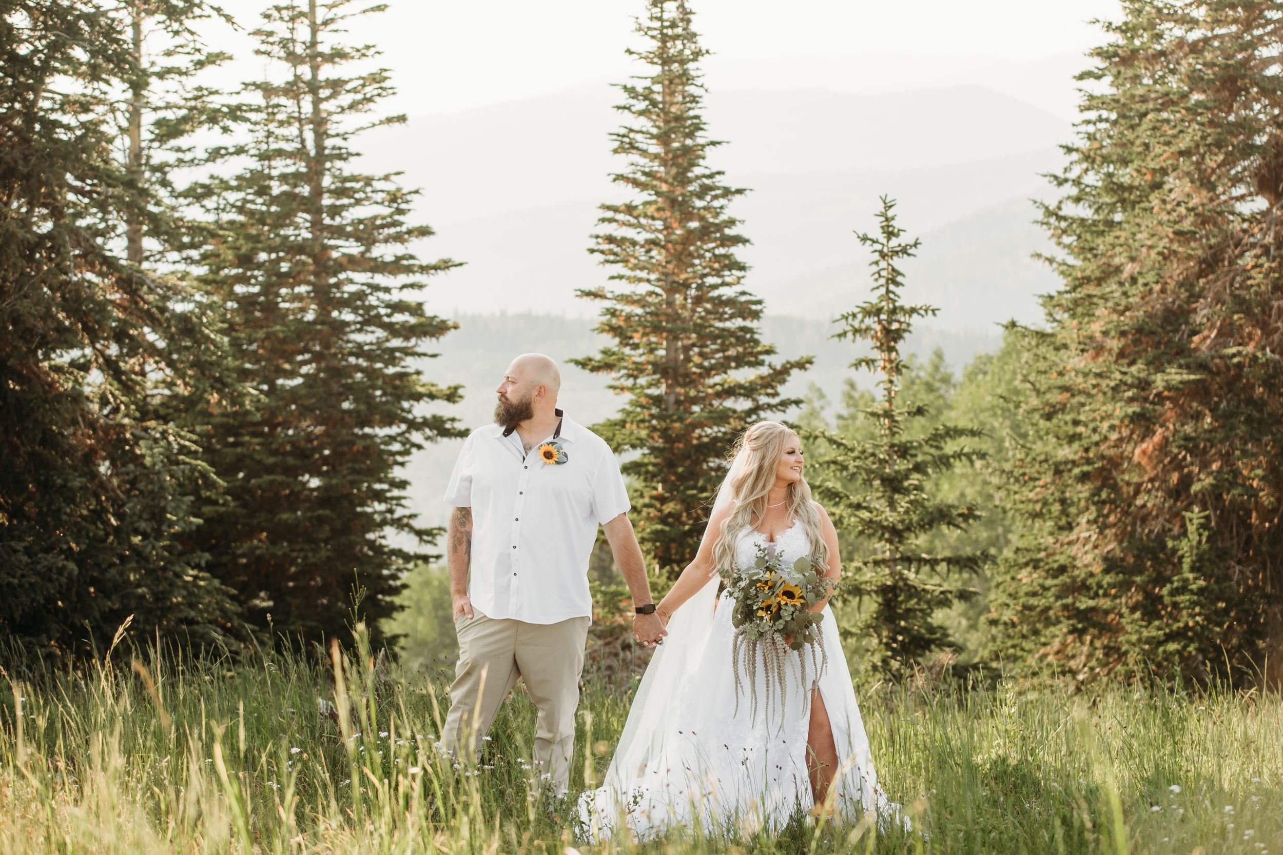Newlyweds walking hand in hand through mountain meadow with evergreen trees.