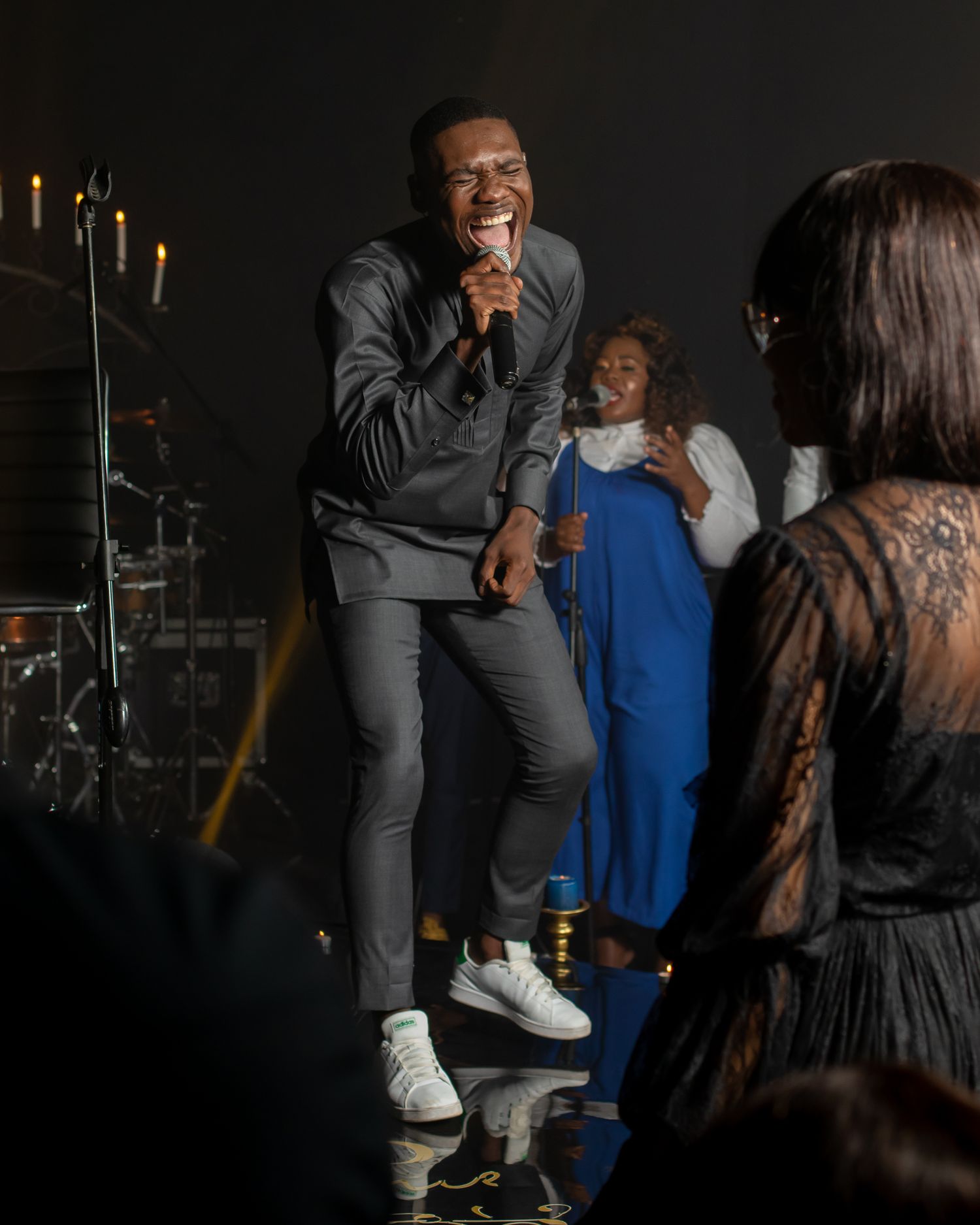 Performer in grey outfit sings on stage with band members visible in background.