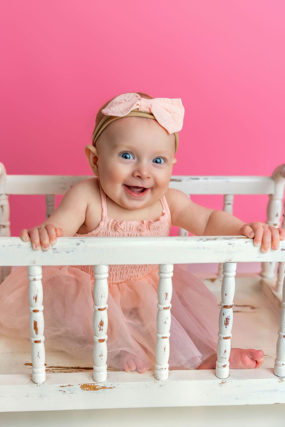 Baby wearing pink dress and bow headband smiles while standing in white vintage crib against pink background.