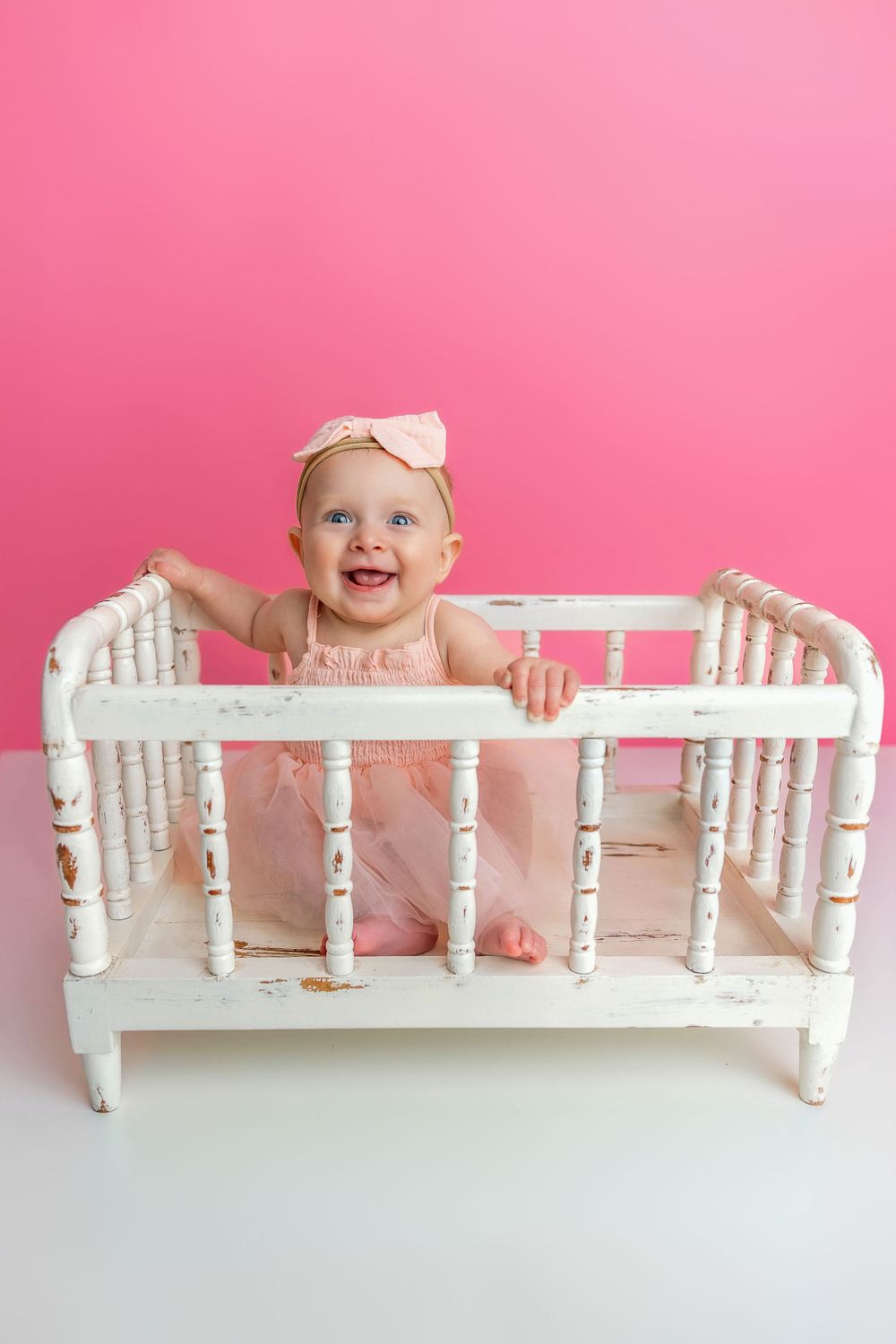 Baby in pink dress stands in distressed white vintage crib against pink backdrop, smiling joyfully.
