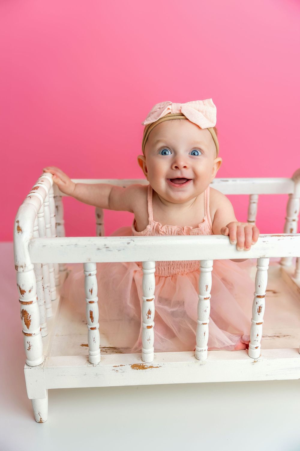 A baby in a pink dress stands in a vintage white crib against a bright pink background.