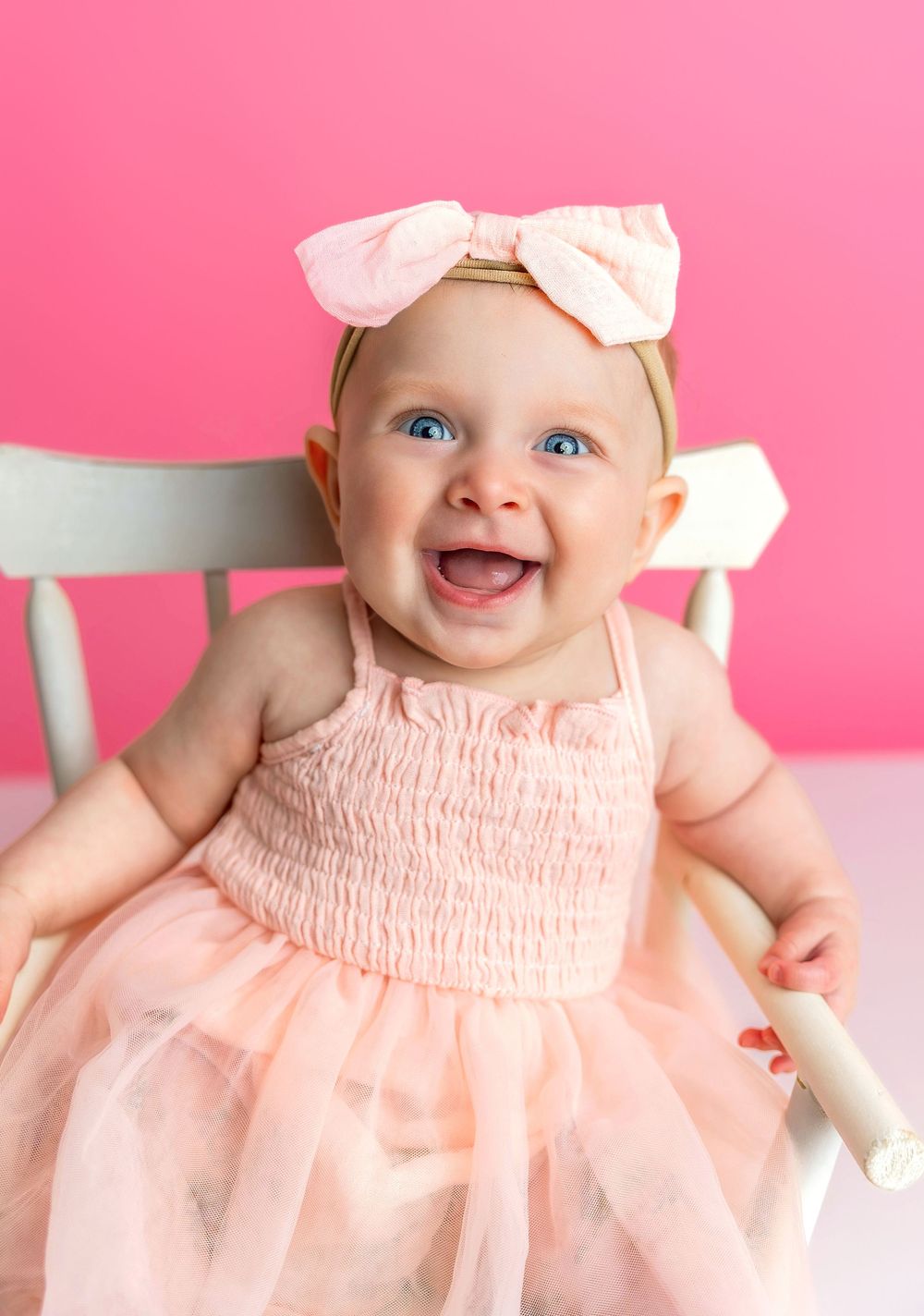 Happy baby wearing pink tulle dress and bow headband sitting in white chair against pink background.