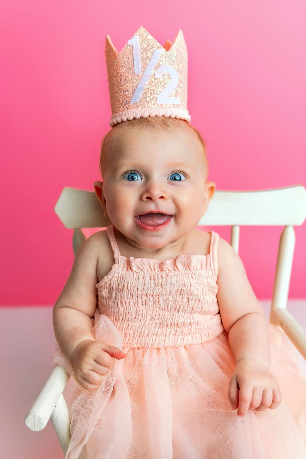 Baby wearing pink tulle dress and sparkly crown sits smiling in white chair against pink background.