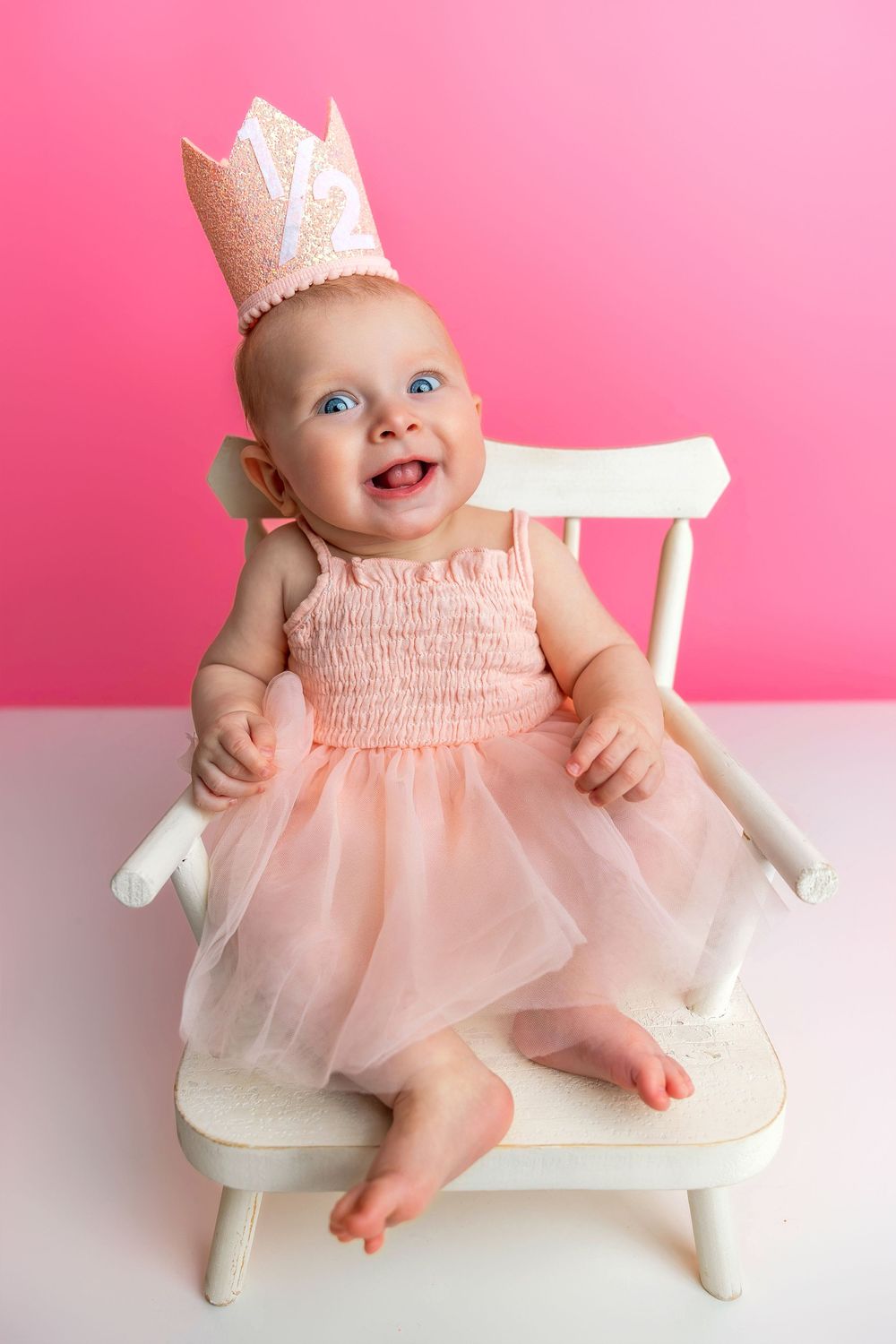 Baby wearing pink tulle dress and glitter crown sits happily on white wooden chair against pink background.