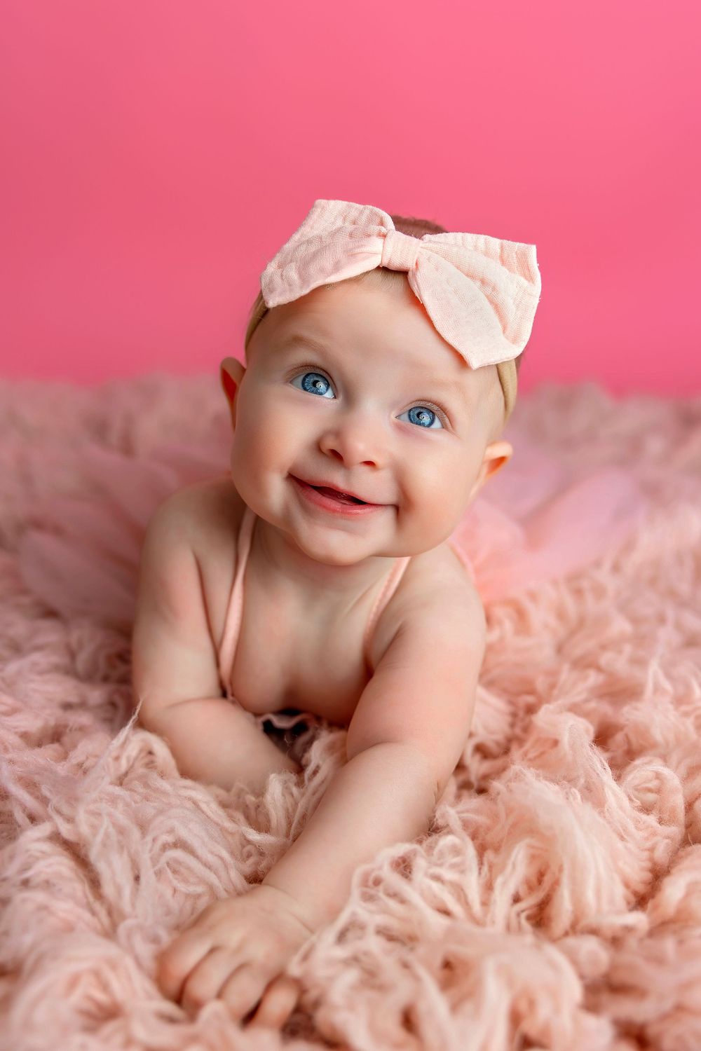 Smiling baby wearing a pink bow headband lying on soft pink textured blanket against pink backdrop.