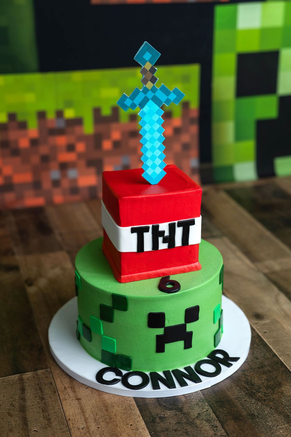Two-tier Minecraft birthday cake with Creeper and TNT design, diamond sword topper, reading Connor age 6.
