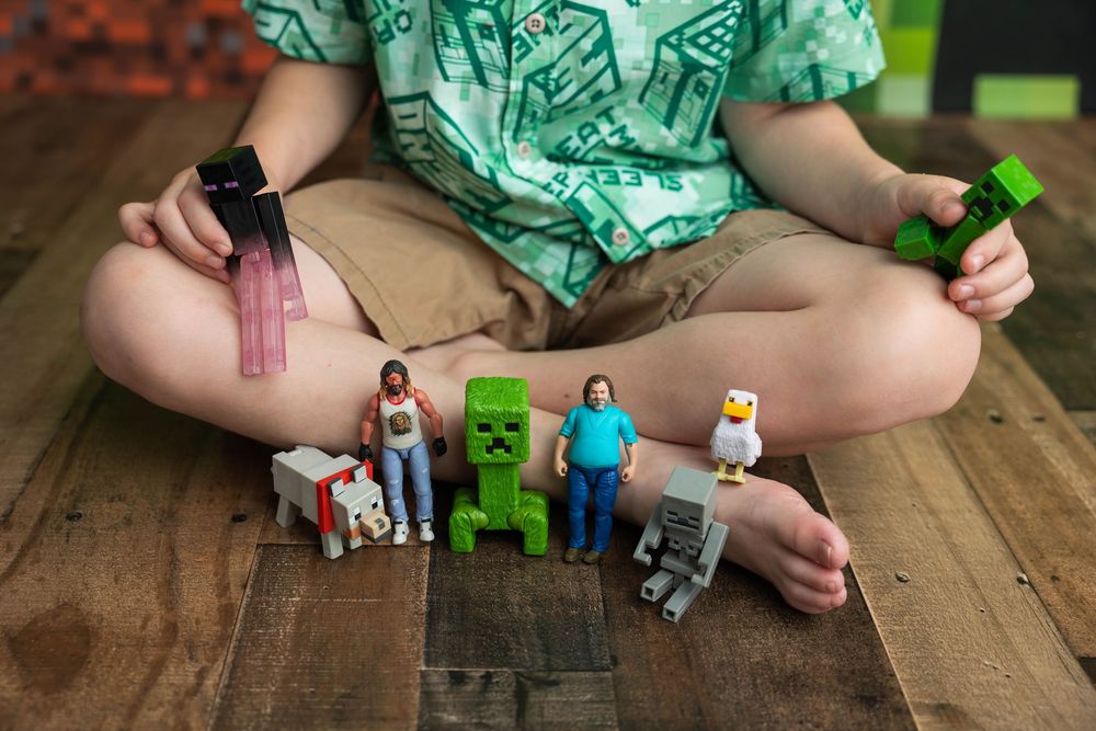 Child sitting cross-legged on wood floor holding Minecraft toys including a Creeper figure and other characters.