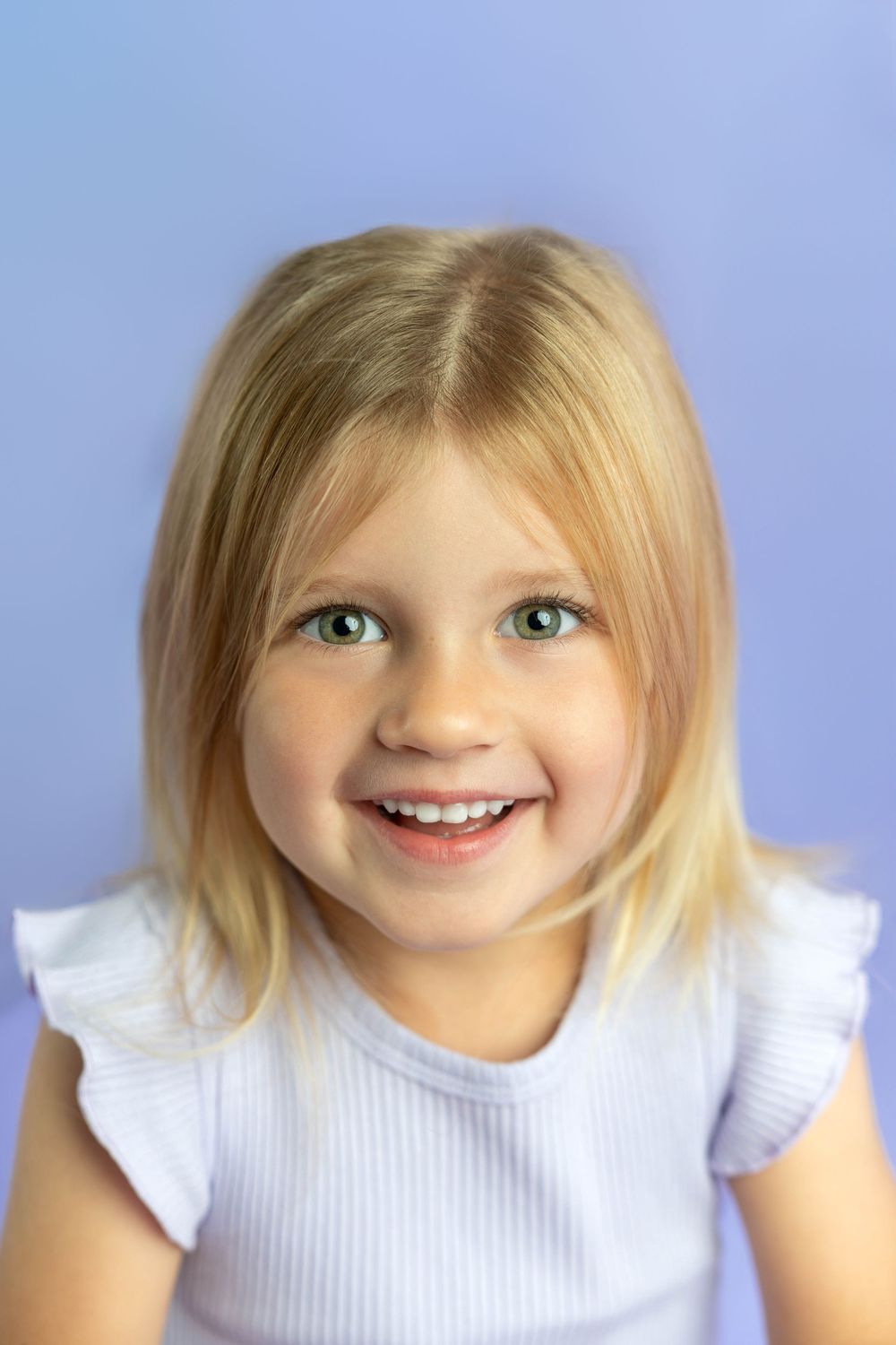 Smiling young girl with blonde hair and green eyes wearing a white ruffle top against a purple background.