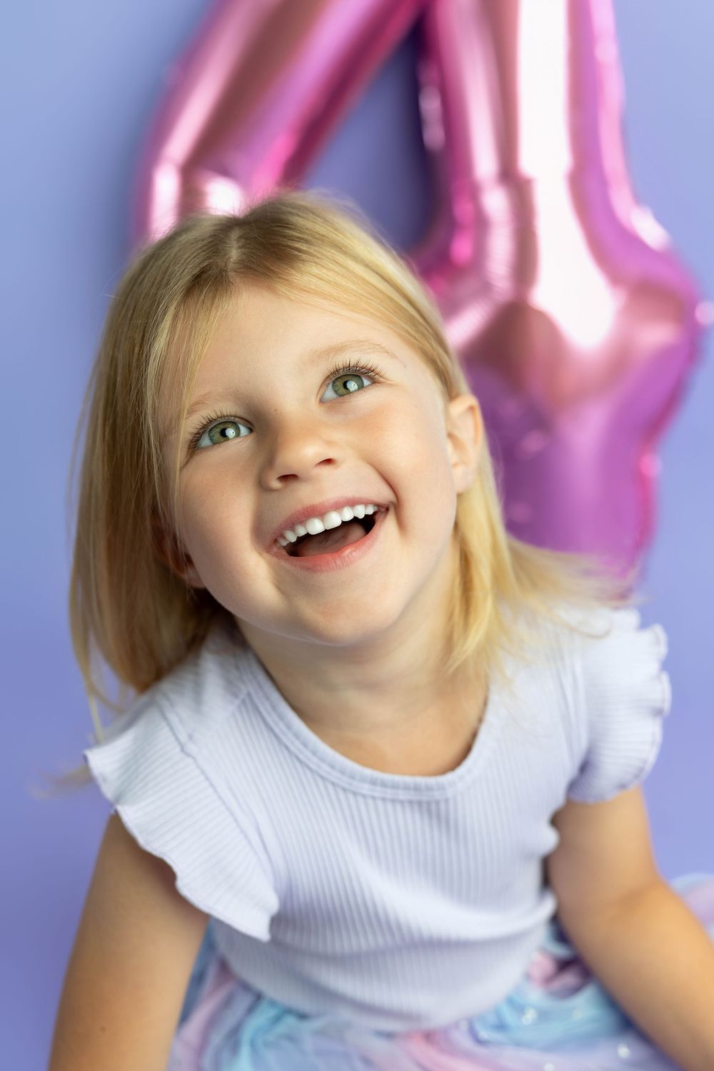 Happy blonde girl laughing in front of a pink number 4 balloon on a purple background.
