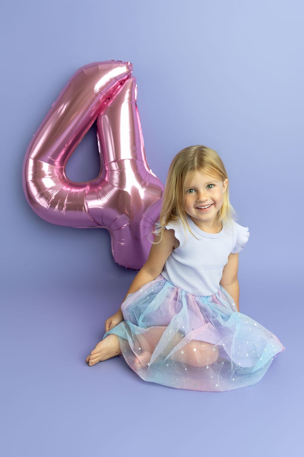 A smiling blonde girl in a tutu sits beside a pink number 4 balloon on a purple background, celebrating her 4th birthday.