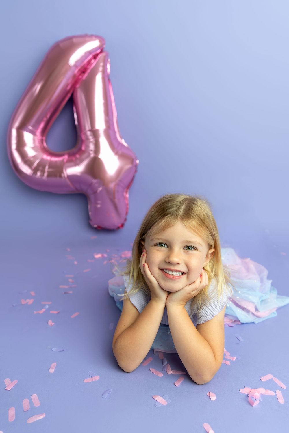 Smiling blonde girl lying on purple floor with pink number 4 balloon and confetti celebrating her 4th birthday.