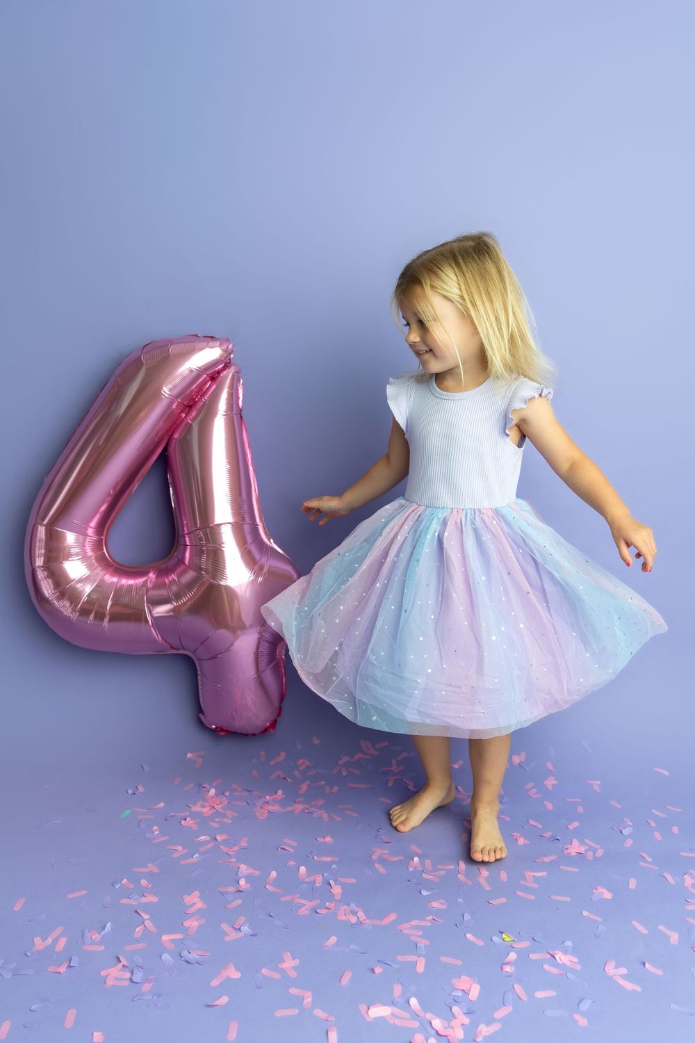 A little girl in a rainbow tutu dress celebrates her 4th birthday with a pink number 4 balloon and confetti.
