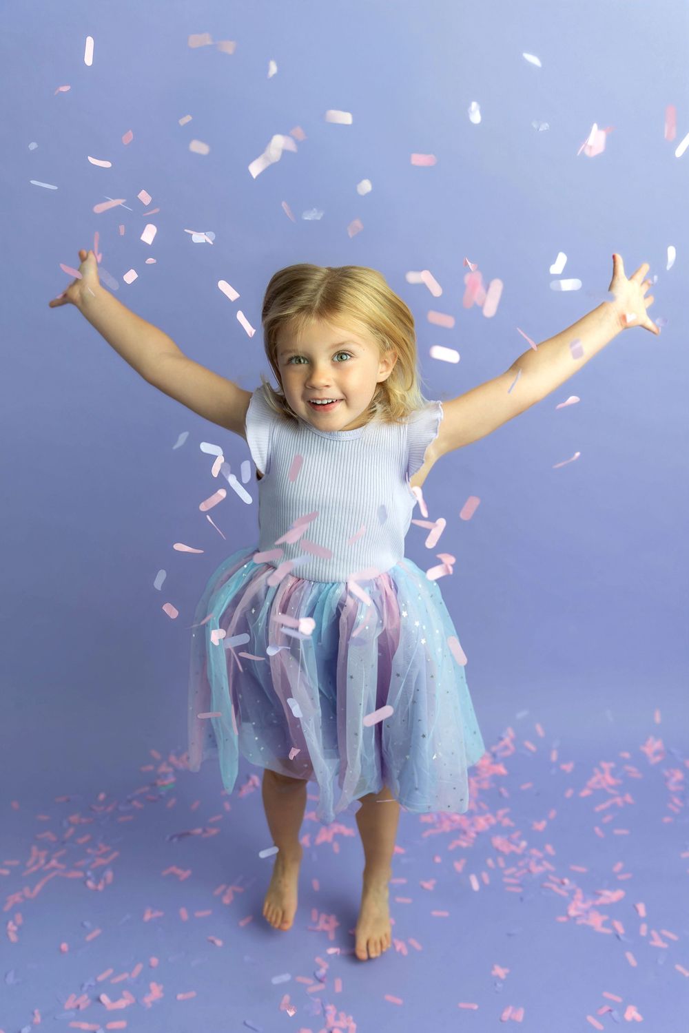A joyful young girl in a blue tutu dress celebrates with arms raised as pink confetti falls around her.