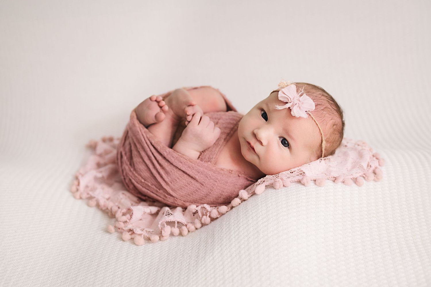 Sleeping newborn baby wrapped in pink fabric with matching headband on white backdrop.
