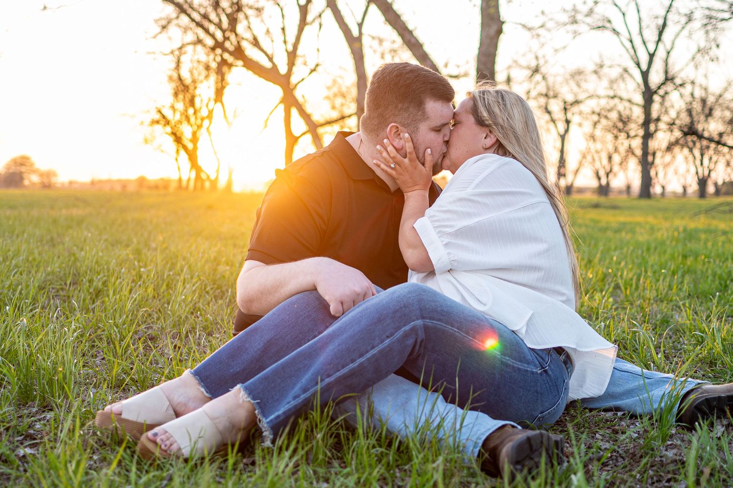 Couple kissing while sitting in a grassy field at sunset with bare trees in the background.