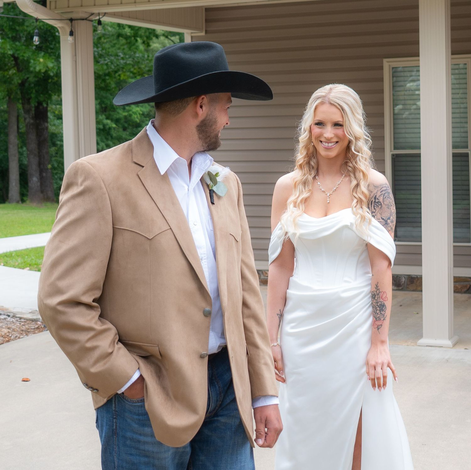 Groom in tan blazer and black cowboy hat gazes at tattooed bride in white wedding dress outdoors.
