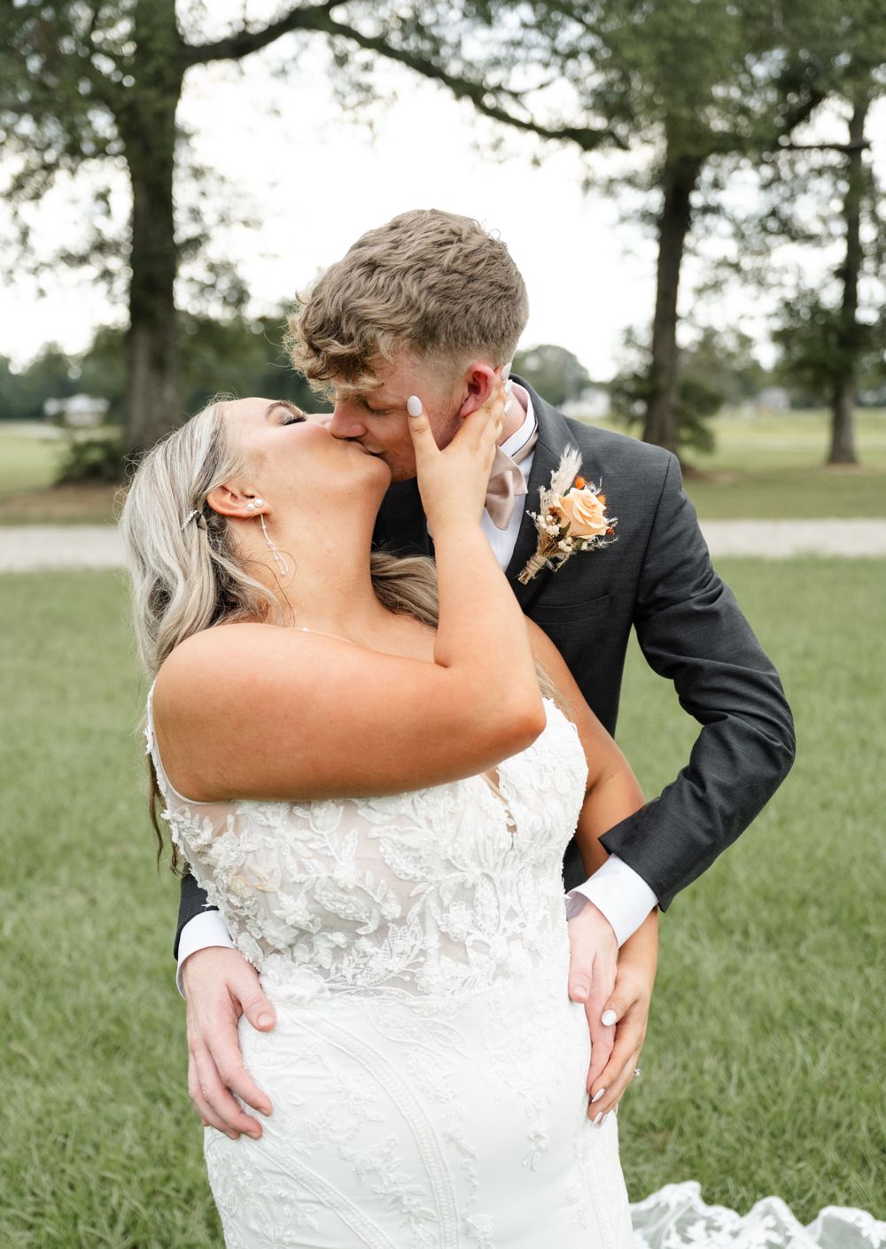 A romantic moment between newlyweds sharing a passionate kiss outdoors in a park surrounded by trees.