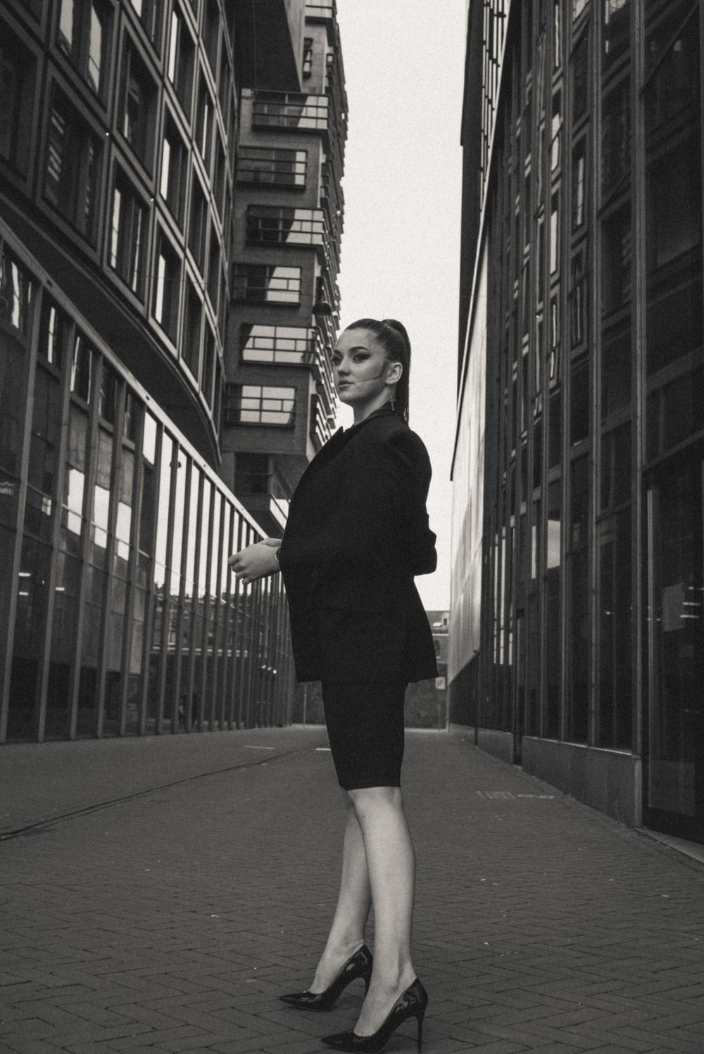 Figure in black dress and heels poses against urban architecture in dramatic low angle shot.
