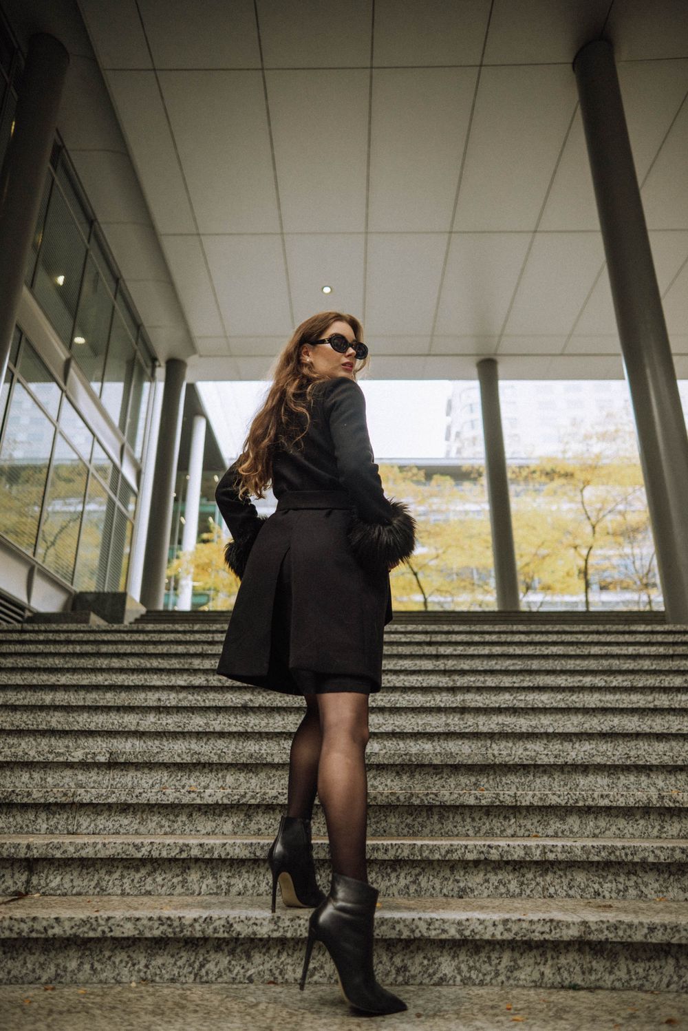 Person in black outfit and sunglasses posing on concrete steps with modern architecture.