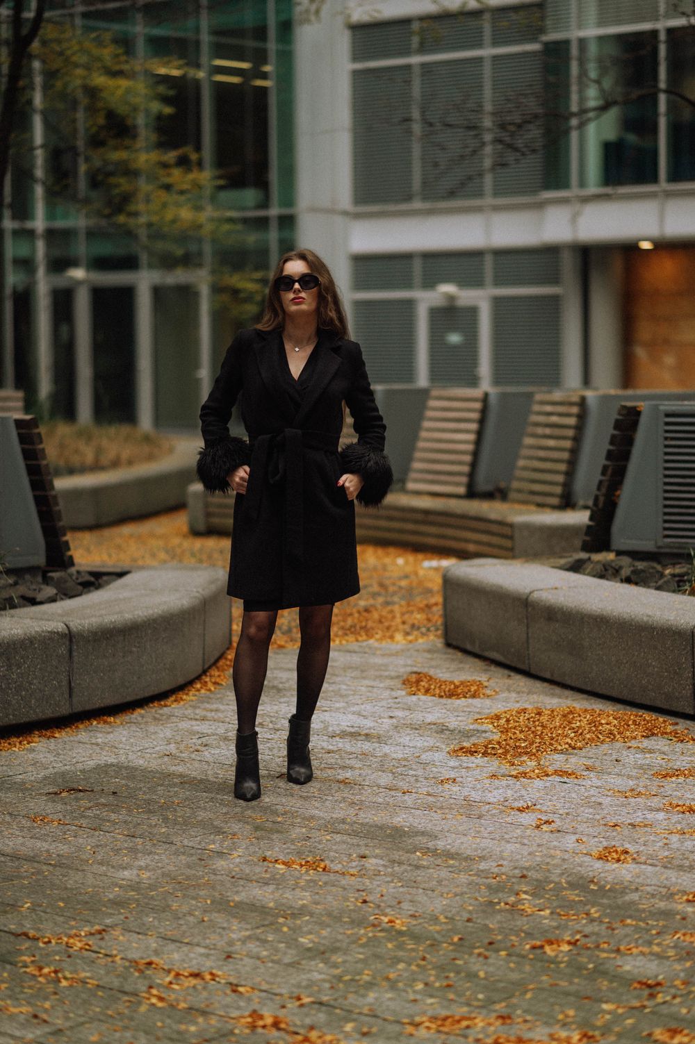 Figure in dark clothing walking on autumn path with fallen leaves near modern building.