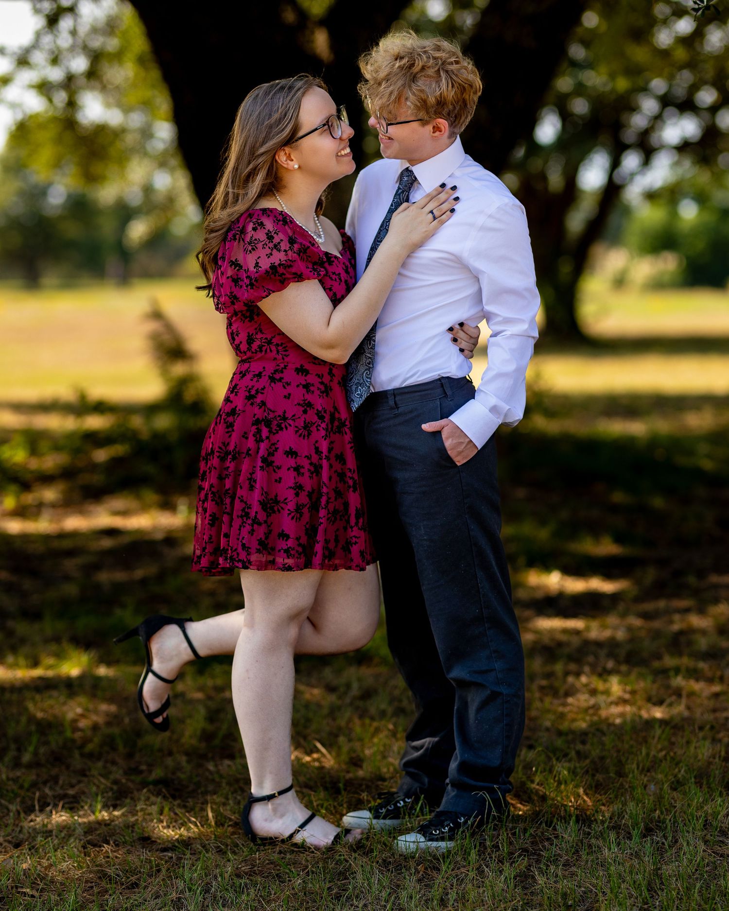 Romantic couple embraces outdoors as woman in burgundy dress lifts her leg while standing under a shady tree.