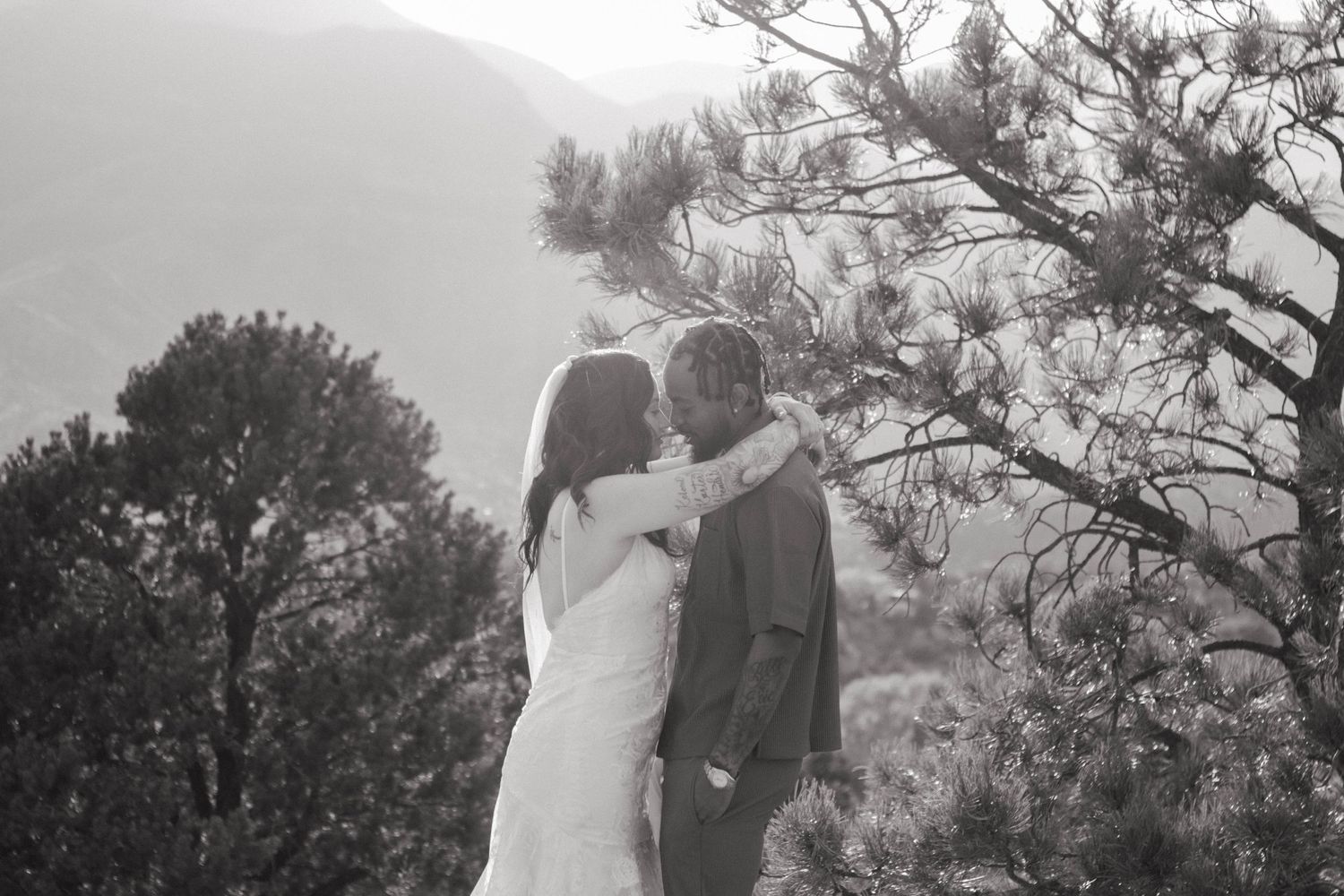 A romantic couple in a wedding dress and suit embrace outdoors with misty mountains and pine trees in the background. Elopement Colorado