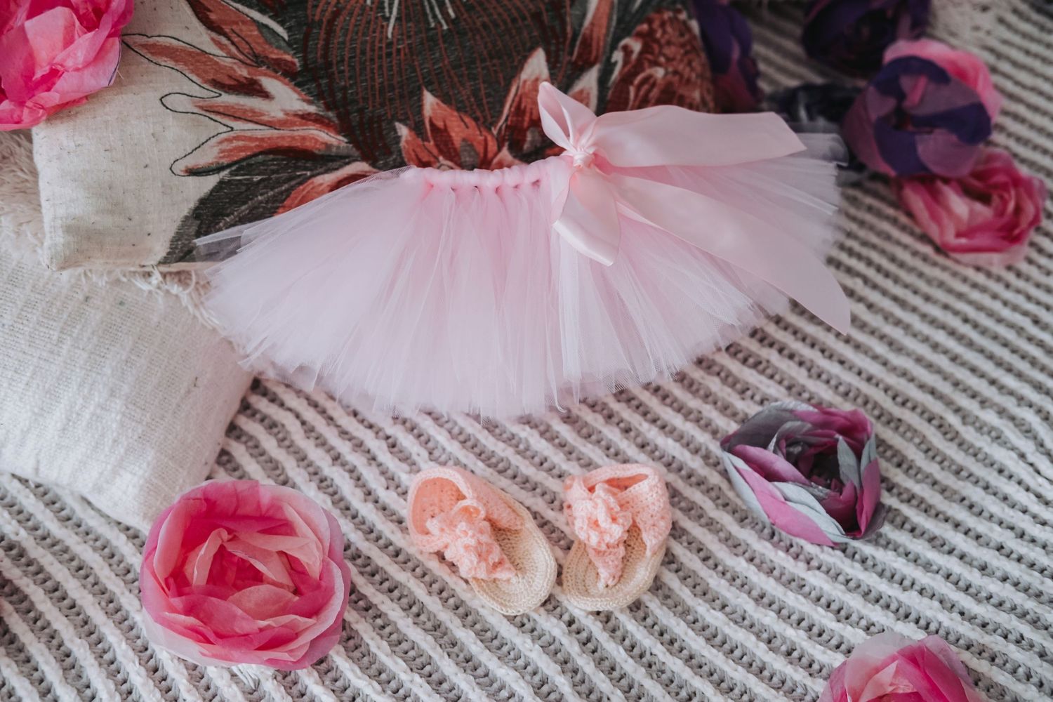 Pink baby tutu skirt with floral shoes and roses arranged on striped blanket.