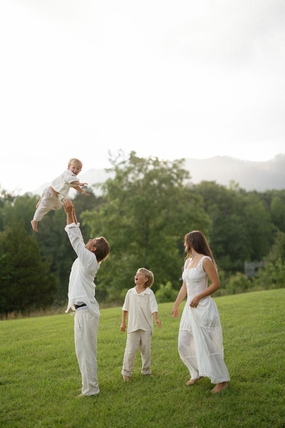A sequence of images showing a family in white clothing playing and lifting a child in a grassy field at sunset.