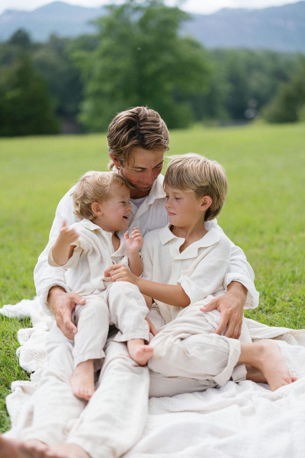 Three connected images show a tender family moment on a white blanket in a sunny meadow with mountains in the background.