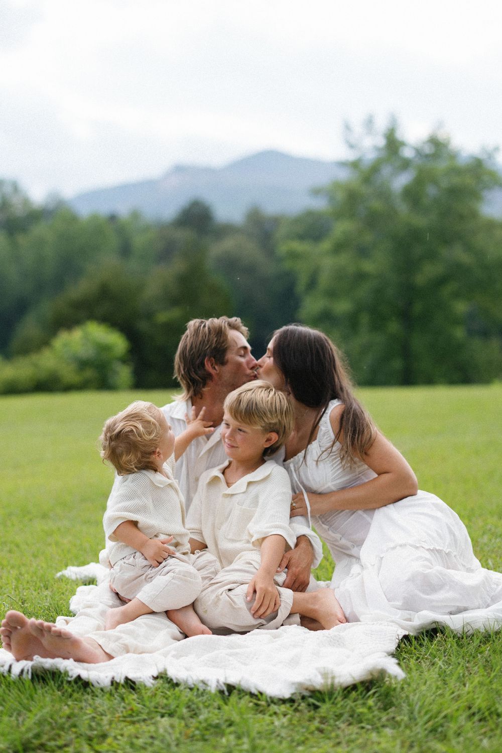 Family enjoying tender moments together while relaxing on white blanket in green meadow with mountains in background.