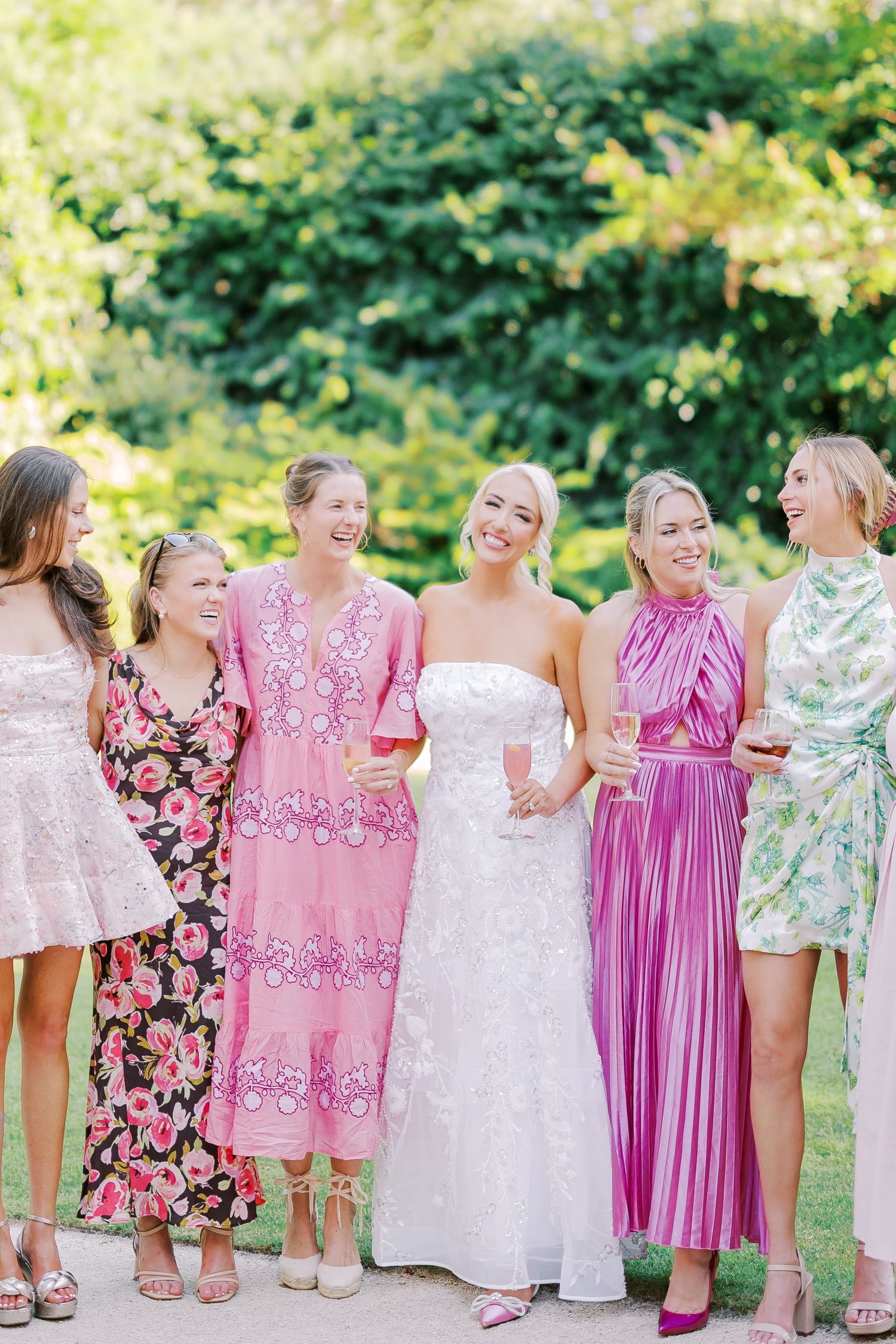 A line of young girls in floral and pink dresses stand with their backs turned at an outdoor summer event.